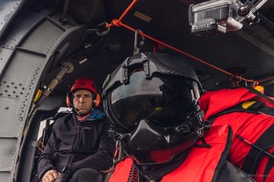 Mężczyzna w czarnym hełmie w pełni zakrywającym twarz i czerwono-czarnym kombinezonie na pokładzie lecącego śmigłowca, za nim kolejny mężczyzna w czerwonym hełmie i słuchawkach oraz granatowej bluzie.