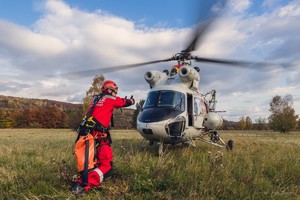 Mężczyzna w czerwonym kombinezonie z napisem STRAŻ na plecach w przyklęku z rękami wyciągniętymi przed siebie i kciukami ku górze daje znak pilotowi śmigłowca, który właśnie wylądował.