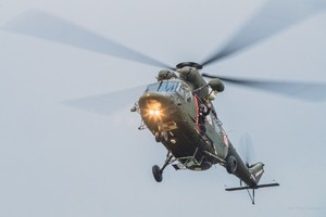 Mężczyzna wychyla się z pokładu ciemno-zielonego helikoptera dając rękami znaki, śmigłowiec ma zapalone lampy na dziobie.