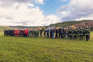 Zbiórka uczestników ćwiczeń służb ratowniczych na trawiastym lądowisku, za nimi policyjny śmigłowiec Black Hawk.