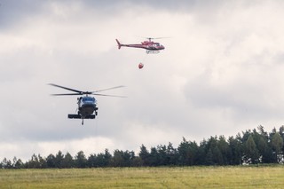 Policyjny śmigłowiec Black Hawk nisko nad lądowiskiem, Powyżej inny śmigłowiec z podwieszonym zbiornikiem Bambi Bucket.