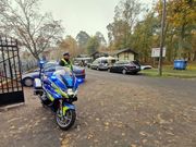 na pierwszym planie policyjny motocykl, za nim stoi umundurowany policjant przed cmentarną bramą