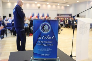 zamazany widok na policjanta, który stoi przed słuchaczami podczas konferencji, z przodu widać wyraźnie statuetkę z napisem: 30 lat negocjacji policyjnych, &quot;Ten kto ratuje życie człowieka, ratuje cały świat&quot; T.Keneally. 2022 rok.