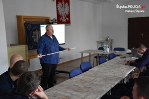 Prowadzący szkolenie trzyma w rękach laskę osoby niewidomej. Przy stołach siedzą policjanci