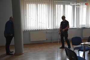 Policjant z zasłoniętymi oczyma porusza się przy pomocy laski. Za filarem stoi mężczyzna