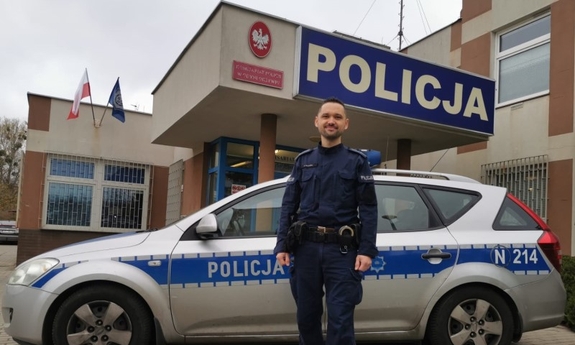 umundurowany policjant stoi przy radiowozie, w tle budynek jednostki Policji