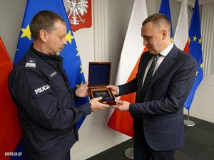 oficer Policji wręcza mężczyźnie w garniturze pamiątkową tabliczkę. Wokół widać stojące w stojakach flagi