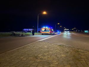 miejsce zdarzenia drogowego, na ziemi leży motorower, a obok niego stoi umundurowany policjant. W tle na ulicy stoi karetka pogotowia ratunkowego