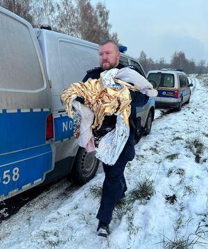 policjant niesie na rękach dziecko owinięte w koc termiczny,  boku widać radiowóz policyjny i auto straży miejskiej