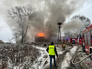 wozy strażackie przed płonącym domem i idący drogą w kierunku pożaru policjant