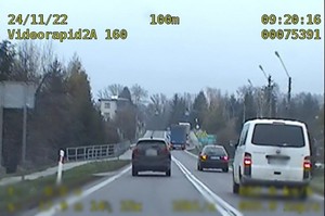 Ciemny samochód wyprzedzający lewym pasem białego busa i jadący przed nim czarny samochód osobowy. W tle naczepy samochodów ciężarowych, elementy infrastruktury drogowej oraz zabudowania