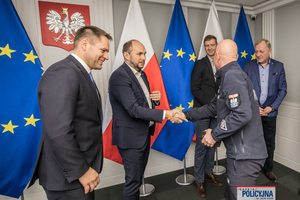 Komendant Główny Policji podaje dłoń przybyłym gościom, z tyłu widać flagi unijne i flagi Polski, a na ścianie godło Polski