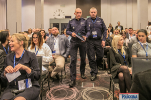 zebrani na sali uczestnicy konferencji. Środkiem idzie dwóch umundurowanych oficerów Policji