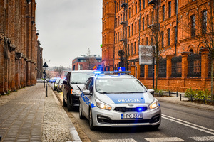 Na drodze sznur pojazdów, pierwszy na drodze stoi radiowóz oznakowany na sygnałach świetlnych, po lewej i prawej stronie widać budynki