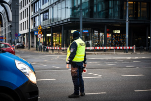skrzyżowanie, na rogu skrzyżowania budynki, na drodze stoi policjant w kamizelce odblaskowej z napisem Policja w ręku trzyma tarczę do kierowania ruchem