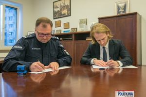 dwaj siedzący przy stole mężczyźni podpisują dokumenty, od lewej umundurowany oficer policji, obok mężczyzna w ubraniu cywilnym