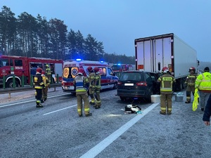 pas jezdni stojący pojazd ciężarowy, pod którego naczepą znajduje się samochód osobowy marki Ford, widać ambulans, wozy straży pożarnej i funkcjonariuszy różnych służb