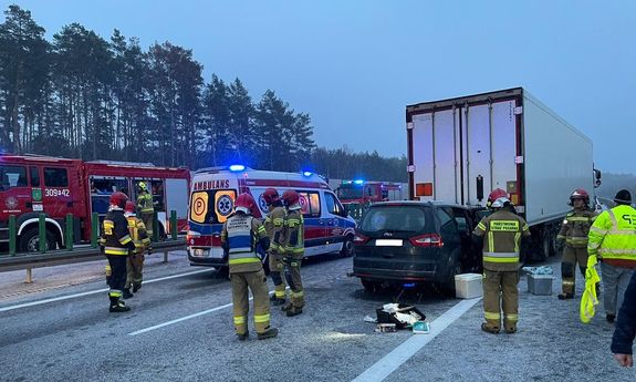 pas jezdni stojący pojazd ciężarowy, pod którego naczepą znajduje się samochód osobowy marki Ford, widać ambulans, wozy straży pożarnej i funkcjonariuszy różnych służb