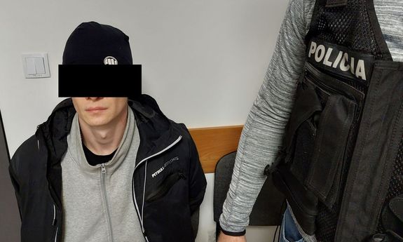 zatrzymany mężczyzna siedzi na krześle, obok niego widać fragment ciała stojącego policjanta, który ma na sobie kamizelkę z napisem Policja