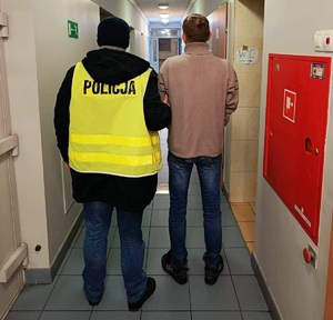 korytarz komisariatu, policjant w żółtej kamizelce odblaskowej prowadzi zatrzymanego mężczyznę