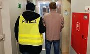korytarz komisariatu, policjant w żółtej kamizelce odblaskowej prowadzi zatrzymanego mężczyznę