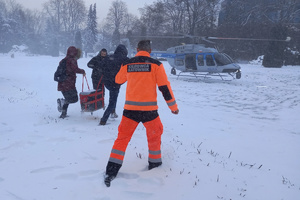 Dwaj mężczyźni niosą pojemnik do transportu organów w kierunku oczekującego śmigłowca. Towarzyszą im dwie inne osoby.