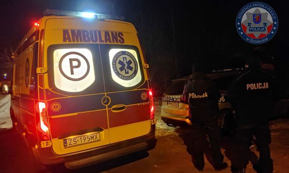 pora nocna. Dwaj policjanci - widok  z tyłu, którzy stoją przy karetce pogotowia. Za nimi widoczny jest fragment policyjnego radiowozu