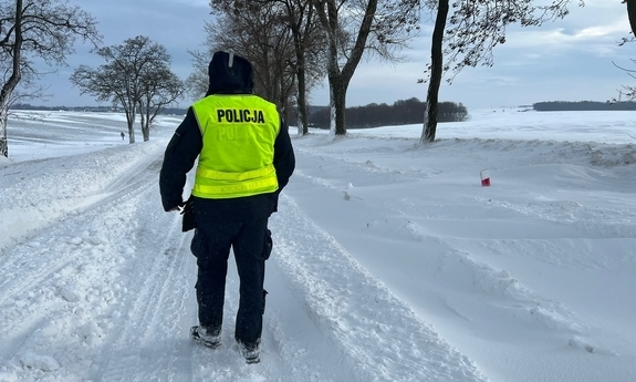 na zdjęciu policjant idący zasypaną śniegiem drogą, na drodze zaspy