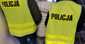 Dwóch policjantów w ubraniach cywilnych, w kamizelkach odblaskowych z napisem Policja prowadzi po schodach zatrzymanego mężczyznę