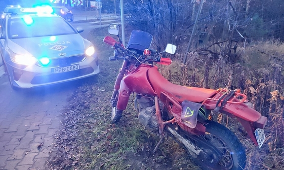 przy drodze stojący czerwony motocykl w tle oznakowany radiowóz z włączonymi światłami błyskowymi