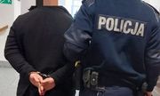 Na korytarzu policjant w granatowym mundurze z napisem policja na plecach trzyma pod rękę osobę w czarnej kurtce i kajdankach założonych na ręce z tyłu - widok z tyłu