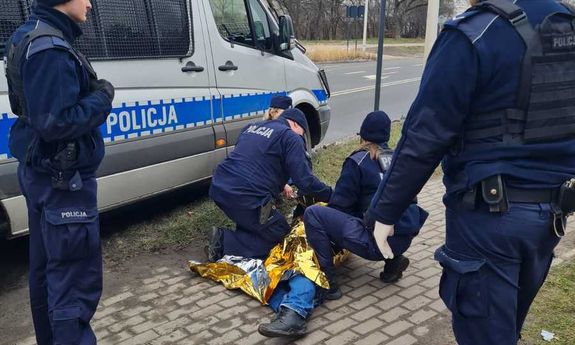 Policjanci udzielają pierwszej pomocy leżącemu na chodniku mężczyźnie. Obok widoczny jest policyjny furgon