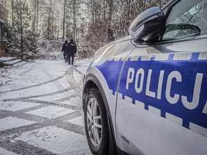policjanci idą droga pokrytą cienką warstwą śniegu, w tle drzewa i zarośla, po prawej stronie fragment radiowozu