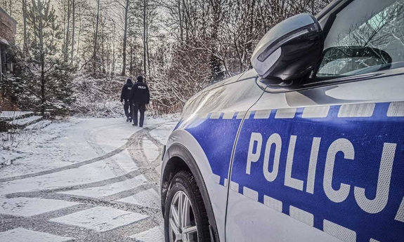 policjanci idą droga pokrytą cienką warstwą śniegu, w tle drzewa i zarośla, po prawej stronie fragment radiowozu