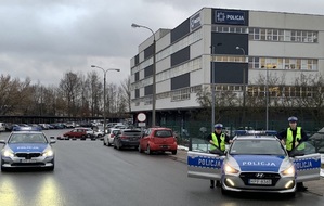 drogą jedzie radiowóz, dwaj policjanci stoją przy radiowozie, w tle budynek komendy Policji