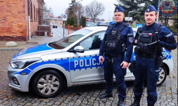 dwóch umundurowanych funkcjonariuszy stoi przy radiowozie