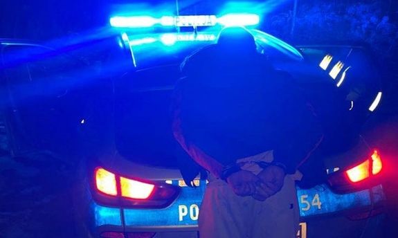 radiowóz oznakowany na sygnałach, mężczyzna z kajdankami założonymi na ręce trzymane z tyłu stoi tyłem do radiowozu