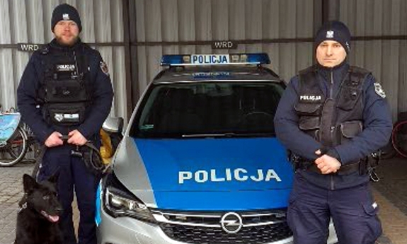 dwóch umundurowanych funkcjonariuszy z psem służbowym stoi przy radiowozie