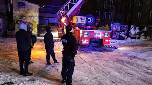 Zdjęcie przedstawia umundurowanego policjanta, który stoi na podwórzu z trzeba innymi ubranymi na ciemno osobami. W tle widać strażaka stojącego przy wozie strażackim, który ma uruchomione niebieskie sygnały świetlne