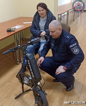 policjant kuca przez elektryczna przystawką do wózka, przy nim na krześle siedzi kobieta