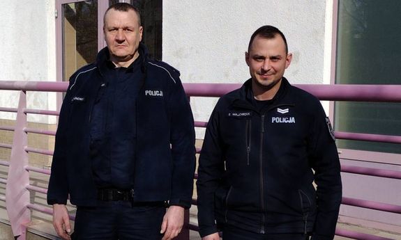 Dwóch umundurowanych policjantów, którzy stoją przodem do obiektywu aparatu. Stoją oni na tle drzwi prowadzących do budynku, nad którym znajduje się czerwoną tabliczka z białym napisem