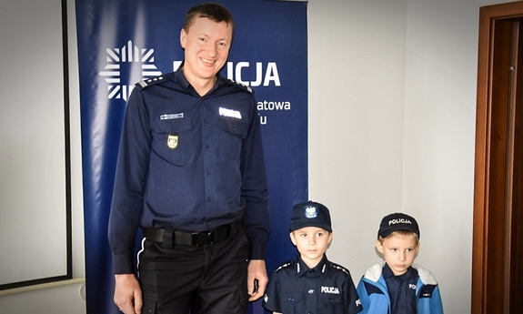 umundurowany funkcjonariusz pozuje do zdjęcia z dwoma chłopcami na tle granatowej ścinaki