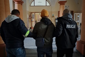 dwaj ubrani po cywilnemu policjanci  w kurtkach z napisem Policja na plecach, prowadzą zatrzymanego