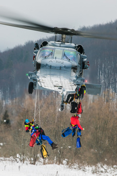 Czterej ratownicy górscy zwisają na linach po bokach lecącego śmigłowca.