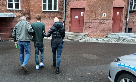 dwoje policjantów kryminalnych prowadzi osobę zatrzymaną przez dziedziniec komendy