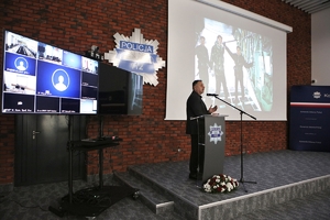 Mężczyzna przemawia do uczestników seminarium stojąc przy mównicy, w tle na ścianie gwiazda policyjna, po lewej stronie zestaw do wideokonferencji, na ekranie po prawej stronie wyświetlana jest prezentacja