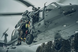 Sfotografowany z ziemi policyjny Black Hawk w zawisie. Z otwartych drzwi maszyny wychylają się zabezpieczeni uprzężami kontrterrorysta, goprowiec oraz crew chief.