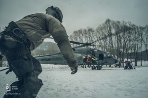 Polowe lądowisko śmigłowców, po ośnieżonej polanie w kierunku zaparkowanego policyjnego Blach Hawka biegnie kontrterrorysta, na pokładzie maszyny uczestnicy ćwiczeń.