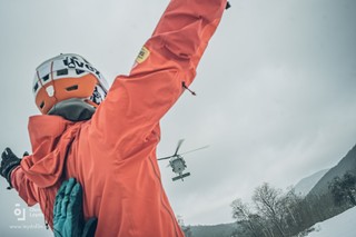 Kadr zza pleców stojącego na ziemi goprowca. Mężczyzna przy pomocy rozłożonych nad głową rak naprowadza lecący w jego kierunku śmigłowiec na lądowisko. Na plecach mężczyzny dłoń asekurująca goprowca czym zabezpiecza ratownika przed możliwością upadku spowodowanego powiewem wiatru zbliżającego się Black Hawka.