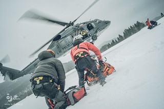 Na pokrytej śniegiem polanie funkcjonariusz BOK oraz ratownik górski klęcząc na śniegu oczekują na lądowanie policyjnego Black Hawka. W odległości kilkunastu metrów przed mężczyznami policyjny śmigłowiec w zawicie. Przed dziobem maszyny dwóch goprowców naprowadzających pilota maszyny na miejsce lądowania.